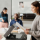 Side view of young Cacuasian manager wearing grey sweater standing in front of printer making copy of document, copy space